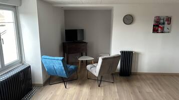 Une pièce avec un plancher en bois, une chaise bleue, une chaise grise, une petite table, un téléviseur posé sur un meuble en bois, un radiateur et une horloge au mur.