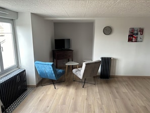Une pièce avec un plancher en bois, une chaise bleue, une chaise grise, une petite table, un téléviseur posé sur un meuble en bois, un radiateur et une horloge au mur.