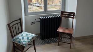 Una habitación con un ventanal grande, dos sillas de madera, un radiador y un cojín estampado.