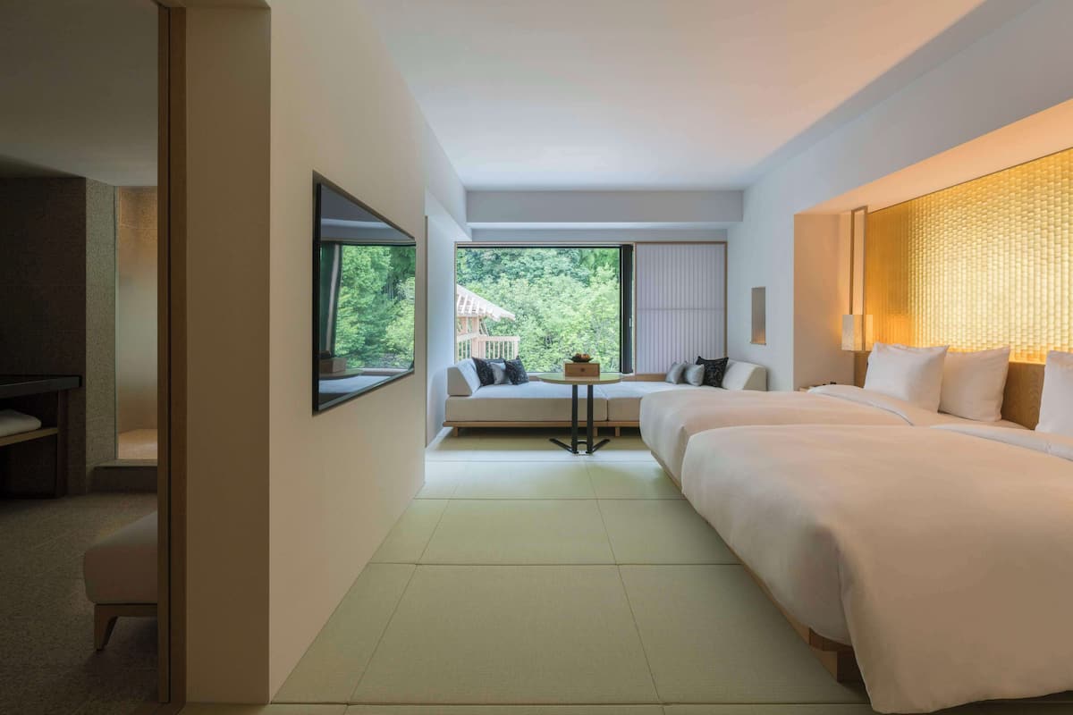 A hotel room with a bed, a sofa, a small table, and a view of greenery outside.
