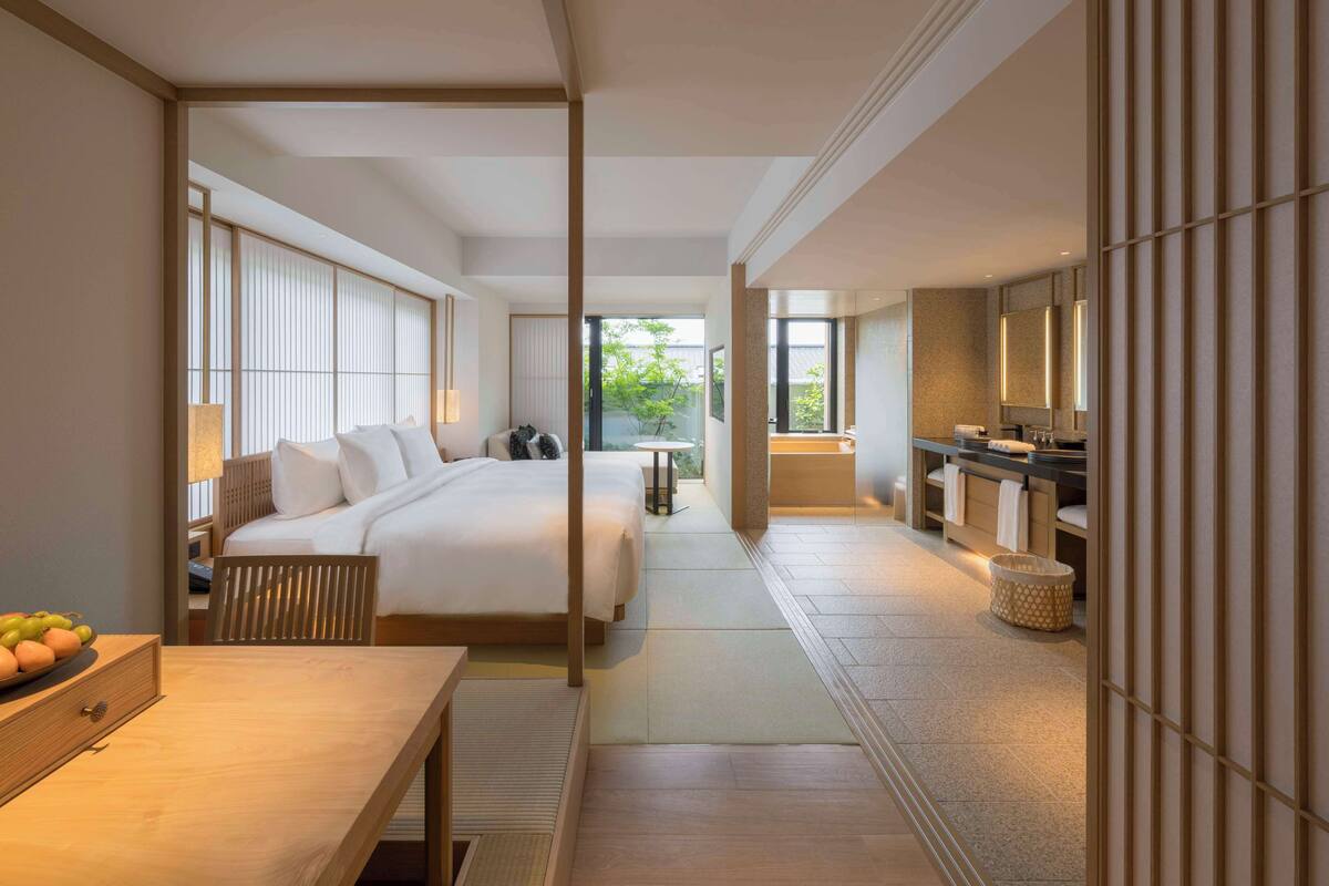 A modern hotel room with a large bed, a desk, a bathroom with a bathtub, and a view of greenery outside.