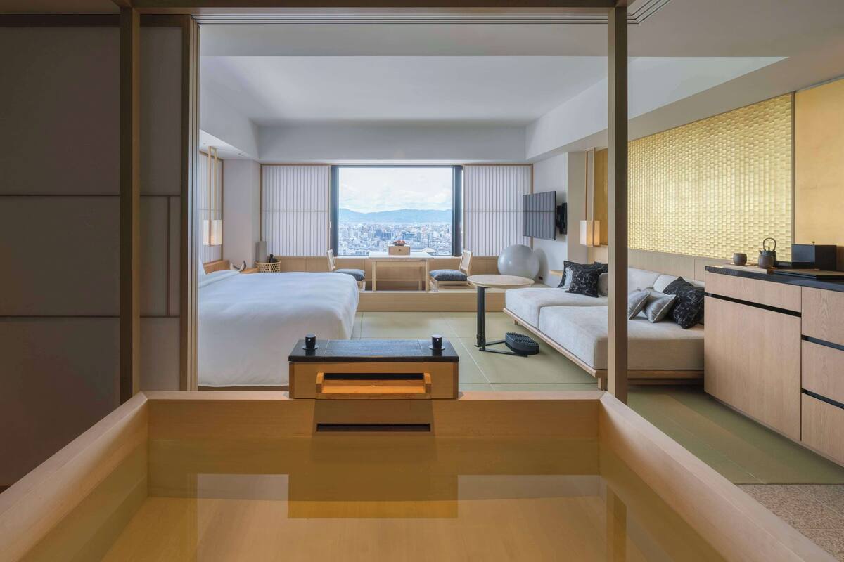 A modern hotel room with a large bed, a desk, a sofa, and a view of the city.