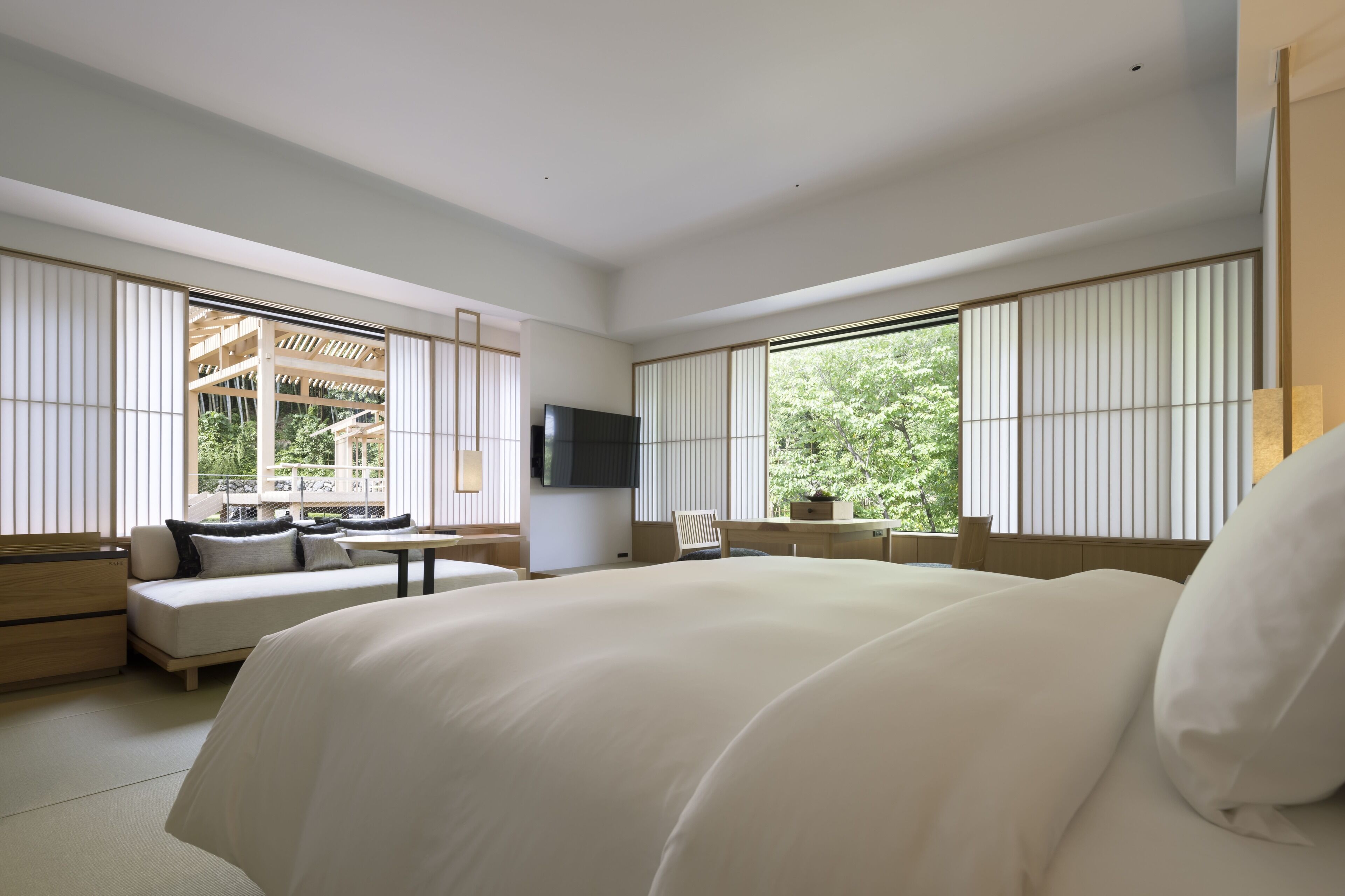 A modern hotel room with a large bed, a sofa, a TV, and large windows with blinds.
