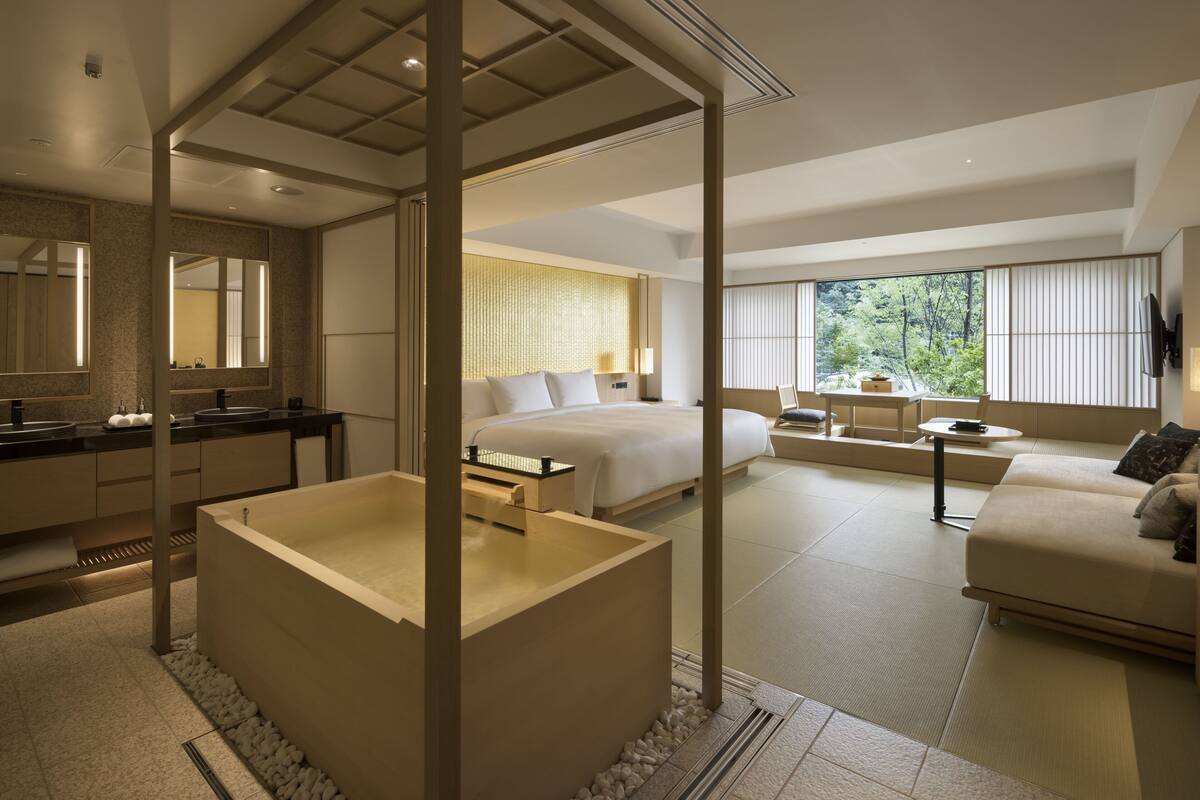 A modern hotel room with a large bed, a sitting area with a sofa and a small table, a bathtub, and a large window offering a view of the outdoors.