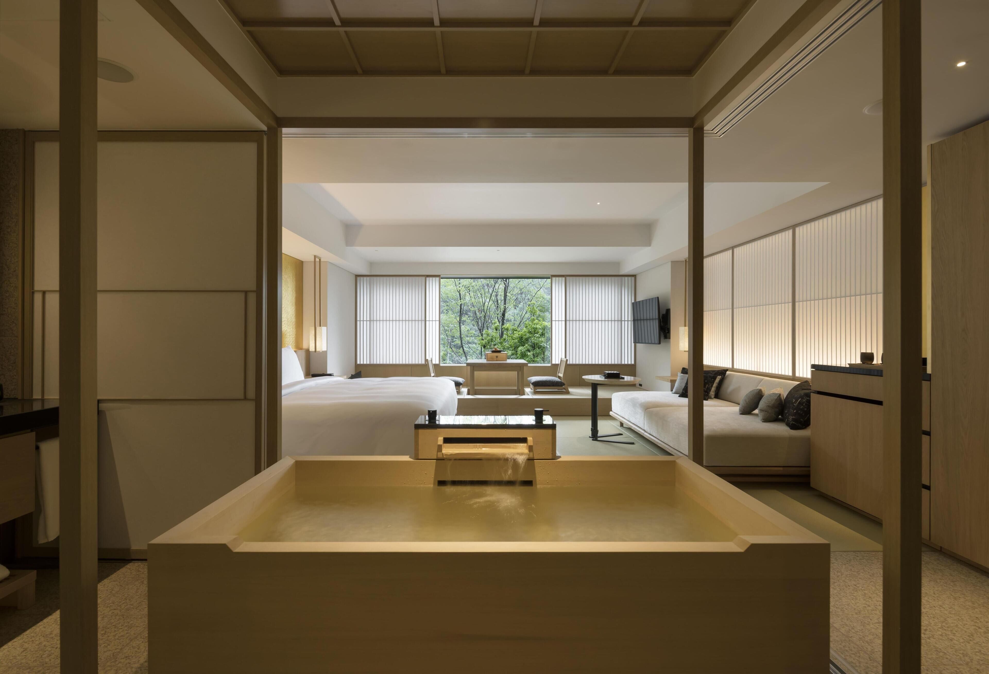 A modern hotel room with a large bathtub, a sofa, a dining table, and a view of greenery outside.