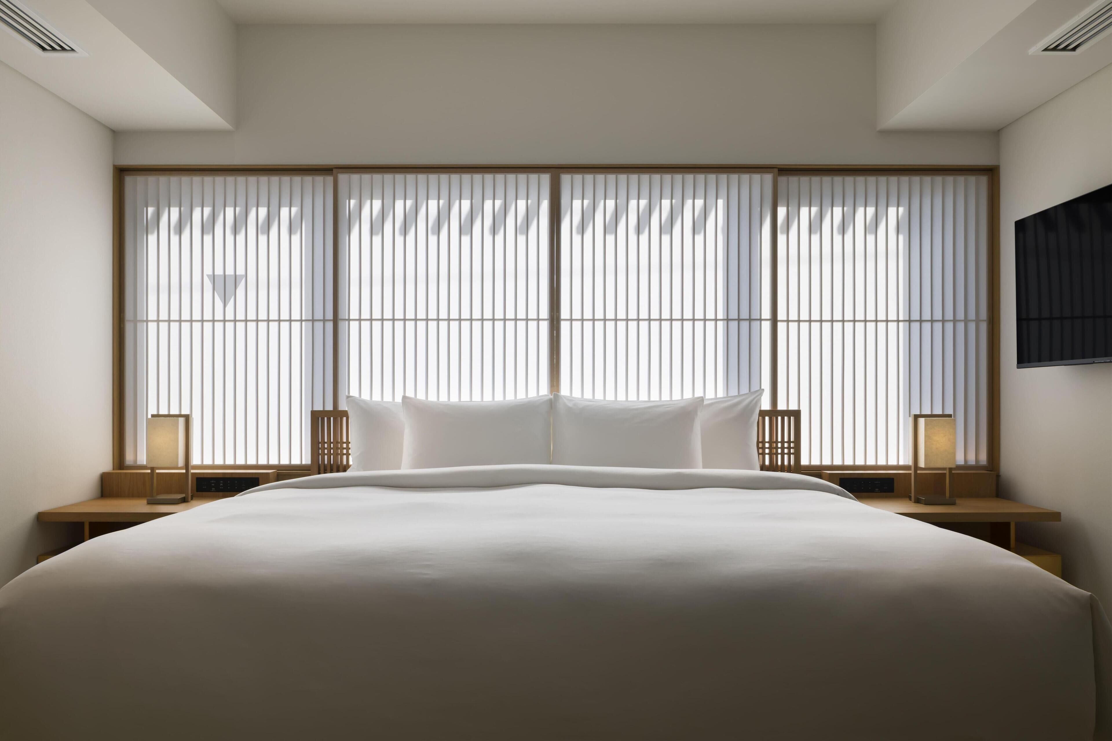 Une chambre d’hôtel moderne avec un grand lit, deux tables de chevet avec des lampes, un téléviseur fixé au mur et une porte coulissante de type shoji.