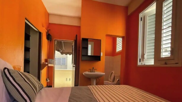 A bedroom with a bed, a bedside table, a window with blinds, a bathroom with a sink and toilet, and a door leading to an outdoor area. - TINY -one room- Apartment / Beach View / Rooftop Terrace  (San Juan)