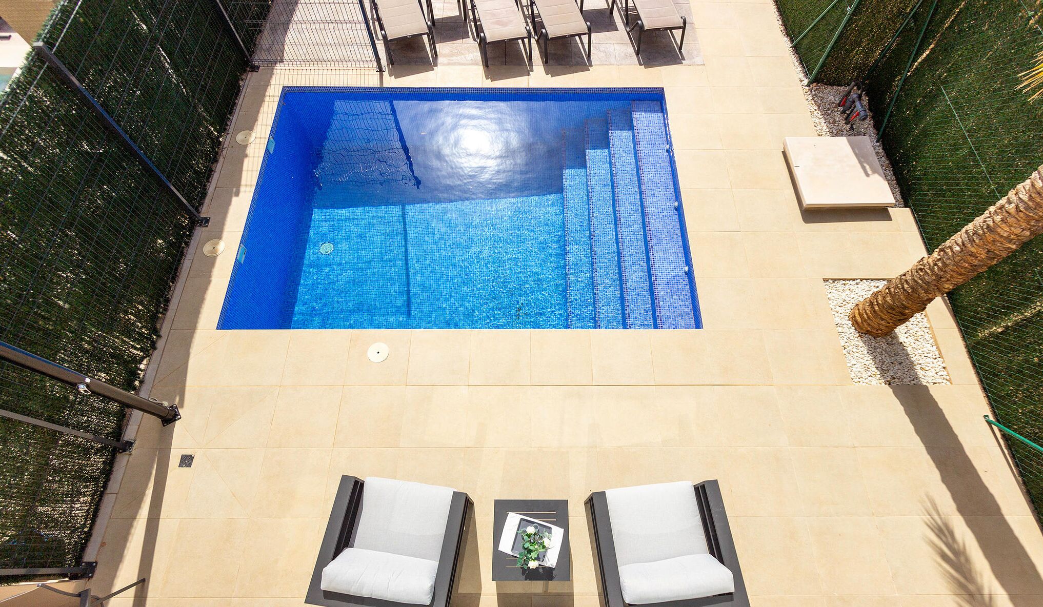 A rectangular pool with a clear blue water surface, surrounded by a tiled deck. There are two lounge chairs with white cushions and a small table with a decorative item.