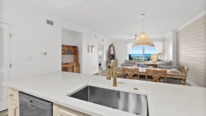 Eine moderne Küche mit weißer Arbeitsplatte, Spüle aus Edelstahl und Armatur in Goldfarbe. Es gibt einen Essbereich mit Holztisch und Stühlen sowie einen Blick auf den Ozean durch große Fenster.