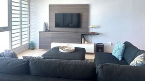 Un salon moderne avec un téléviseur à écran plat fixé sur un mur en bois, un ventilateur de plafond et une grande fenêtre équipée de stores.