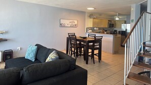 Un salon avec un canapé aux couleurs sombres et un coin repas comprenant une table et des chaises en bois. On aperçoit une cuisine à l’arrière-plan, équipée d’appareils électroménagers modernes.