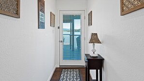 A hallway with a door leading to a balcony with a view of a lake and mountains. There are framed welcome signs and a table with a lamp. - Lakefront, 1 King Bed, dock, mist view, electronic firepit (Lower Lake)