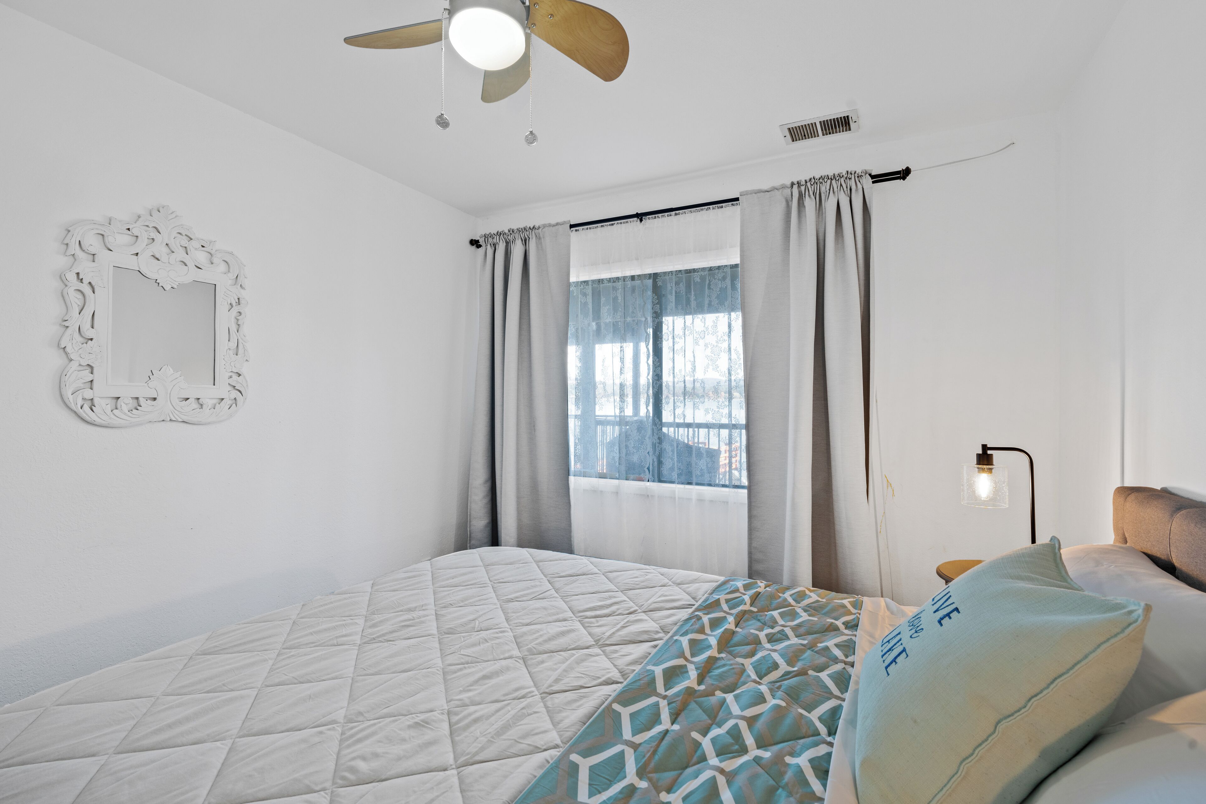 Un dormitorio con cama, ventilador de techo, ventana con cortinas, espejo montado en la pared y lámpara de noche.