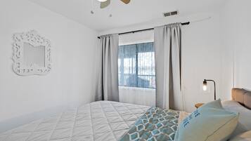 Un dormitorio con cama, ventilador de techo, ventana con cortinas, espejo montado en la pared y lámpara de noche.