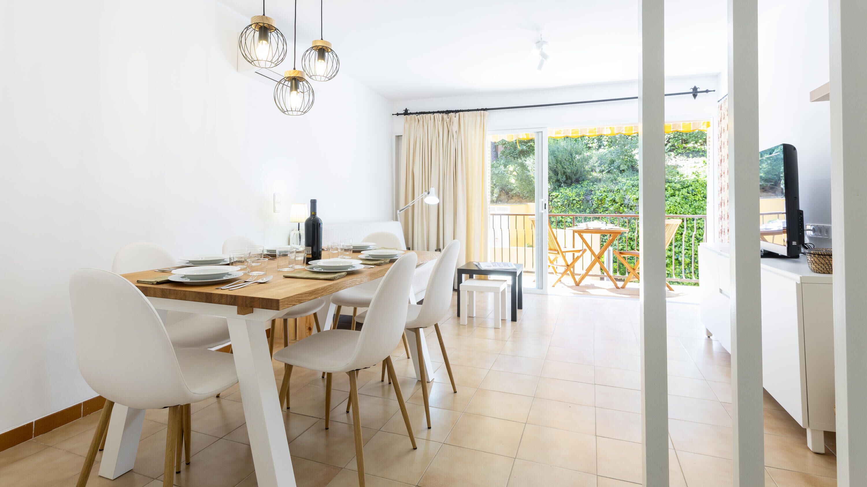 Un coin repas avec une table en bois dressée pour quatre personnes, des chaises blanches et des luminaires suspendus.
