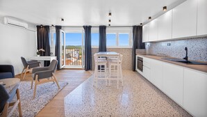Una cocina moderna con armarios blancos, encimera de madera y grifo negro. Cuenta con una zona de comedor con mesa y sillas, y un balcón con vistas a la ciudad.