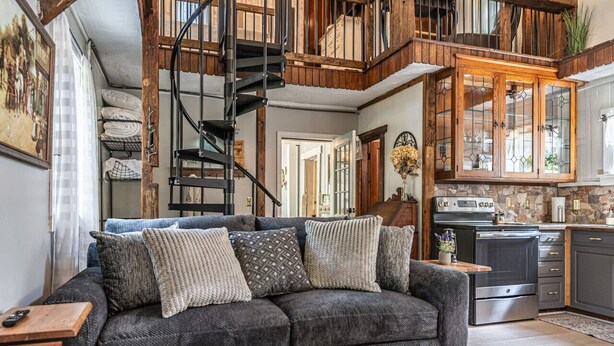 A cozy living room with a grey sofa, wooden accents, a spiral staircase, and a kitchen area with a stove and cabinets.
