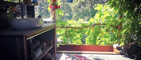 A room with a view of a lush garden, a wooden table with a bowl and a lamp, and a colorful rug on the floor.
