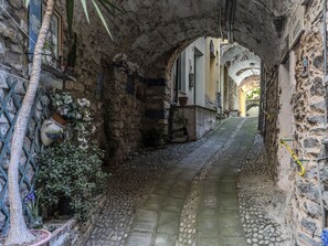 A narrow stone pathway flanked by buildings with arched doorways, leading to a courtyard.