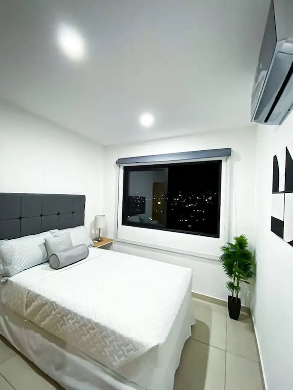 Un dormitorio moderno con una cama grande, una mesita de noche, un ventanal con vistas a luces de ciudad y una planta en maceta.