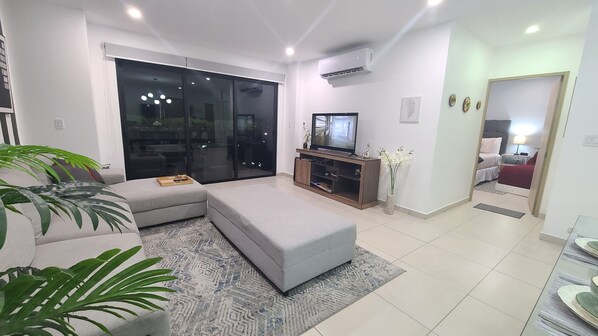 Una sala de estar moderna con un sofá seccional gris, un televisor de pantalla plana sobre un mueble de madera y un amplio ventanal corredizo que da a un balcón.