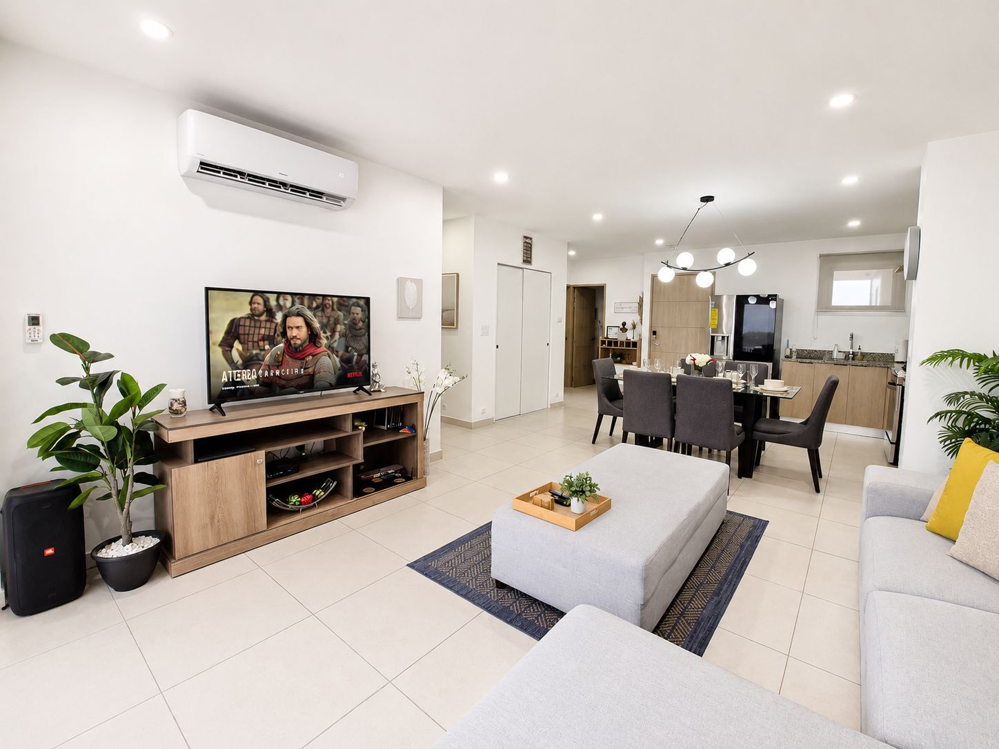 A modern living room with a flat-screen TV, a dining area with a table and chairs, and a kitchen in the background.