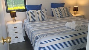 A neatly made bed with striped bedding, two bedside lamps, and a window with a view of greenery. - Broadbeach Tropical Gem with Lux features (Broadbeach Waters)