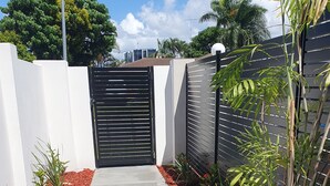 A paved pathway leading to a gated entrance, surrounded by red mulch, white walls, and various plants. - Broadbeach Tropical Gem with Lux features (Broadbeach Waters)