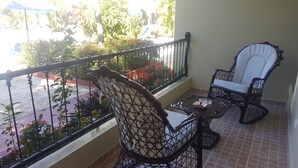 Un balcón con dos sillas de mimbre y una mesita, con vistas a un jardín con flores rojas y una calle.