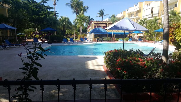 Una piscina con sillones de descanso y sombrillas, rodeada de palmeras y un edificio con balcones.