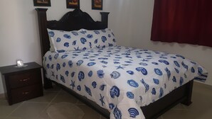 Una cama con una colcha blanca y azul estampada, un cabecero de madera oscura, una mesita de noche pequeña con una lámpara y una cortina roja.