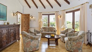 A spacious living room with a high ceiling, wooden beams, a large window with curtains, and a wooden coffee table with fruit. - Navarra by Interhome (Moraira)