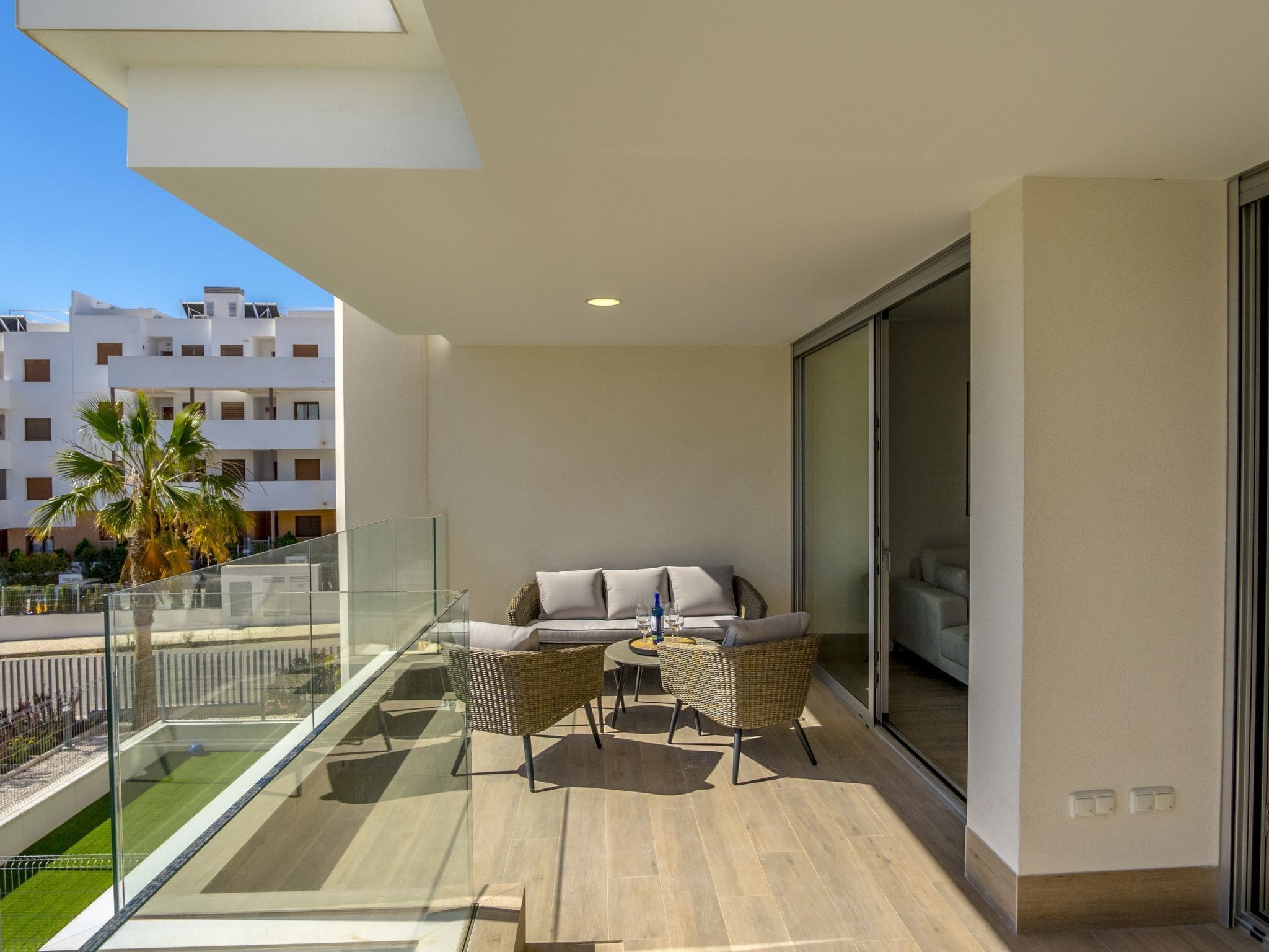 A balcony with a glass railing, a sofa, two wicker chairs, and a small table with a bottle and glasses.