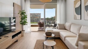 A modern living room with a flat-screen TV, a sofa, a coffee table, and a large window offering a view of a building and greenery.