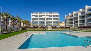 A modern hotel with a swimming pool, palm trees, and a clear blue sky.