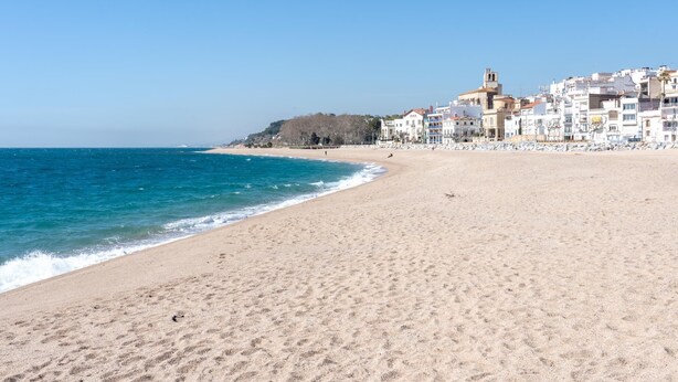 A sandy beach with a clear blue sea, a row of buildings with balconies, and a hill in the background. - Ideal long stay: central, parking and pool (Sant Pol de Mar)