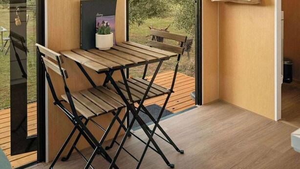 A modern room with a wooden floor, a foldable table, a bench, a hat on the wall, and a bag with a logo.