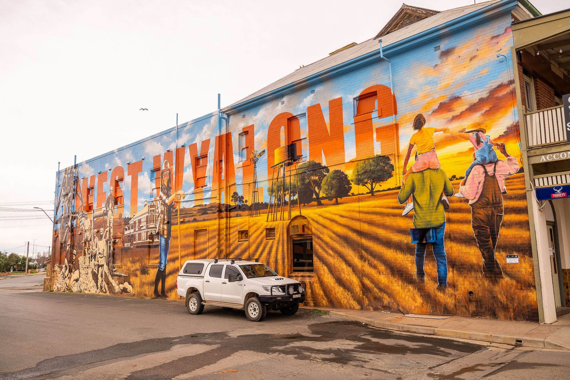 Un mural en un edificio con letras y figuras grandes, una camioneta blanca estacionada y un escaparate con un cartel.