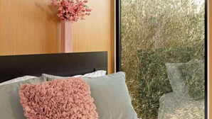 A bedroom with a large window, a bed with a white sheet, a pink fluffy pillow, a vase with pink flowers, and a round wall-mounted light.