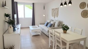 Un salon moderne avec un canapé, une table à manger et des chaises. On y trouve une fenêtre avec des rideaux, une décoration murale en macramé et une lampe.