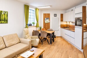 Una sala de estar moderna con un sofá, una mesa de comedor con sillas y una cocina con armarios blancos y horno empotrado.