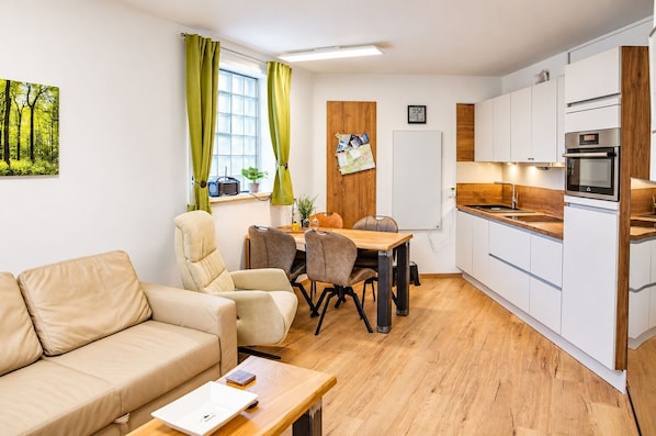 Ein modernes Wohnzimmer mit einer Couch, einem Esstisch mit Stühlen und einer Küchenzeile mit weißen Schränken und einem eingebauten Backofen.