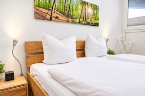 Ein Schlafzimmer mit einem hölzernen Kopfteil, weißer Bettwäsche, einem Sony-Wecker, einem Bild von einem Wald und einem weißen Stuhl.