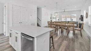 A modern kitchen with white cabinetry, a central island, and a dining area with wooden chairs and a table. - Twente Toes (Navarre)