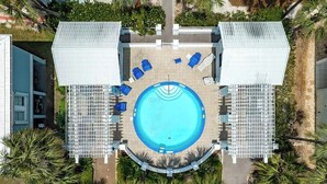 An aerial view of a hotel with a central swimming pool surrounded by lounge chairs and sunbeds, with palm trees and a brick building in the background. - Twente Toes (Navarre)