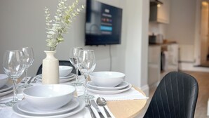 Un coin repas avec une table ronde dressée pour quatre personnes, des assiettes blanches, des verres et des couverts. Un vase avec une plante est posé sur la table. Un téléviseur est fixé au mur au fond de la pièce.
