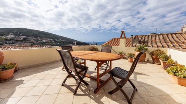 Eine Terrasse mit Holztisch und -stühlen, Blumentöpfen und Blick auf eine Stadt und sanfte Hügel.