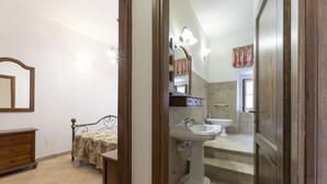Un baño con lavamanos, inodoro y espejo. En la habitación contigua, hay una cama con una colcha estampada.