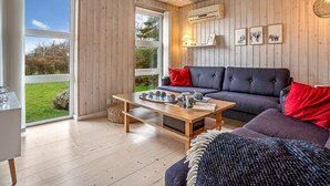 Een moderne woonkamer met houten vloer, een grijze bank, een houten salontafel en uitzicht op de omgeving door grote ramen.