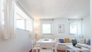 Uma sala de estar luminosa com um sofá branco, uma mesa de jantar com cadeiras e uma mesa de centro. Há duas janelas com cortinas, um abajur e um quadro na parede.